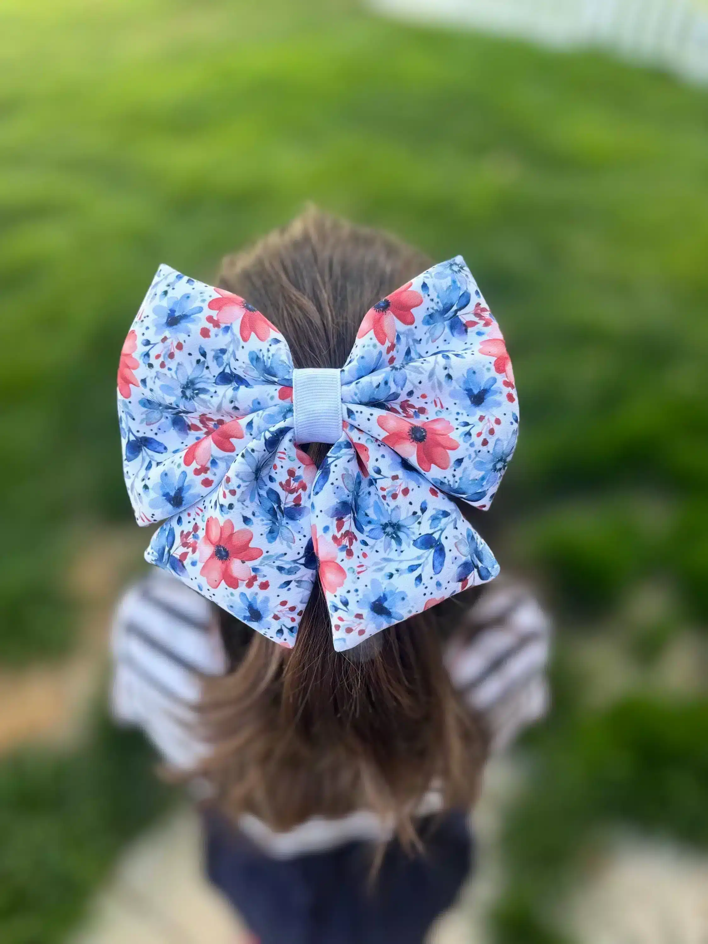 A handmade puffy hair bow with a patriotic floral print, worn by a girl, displayed against a green outdoor background. The bow features red, white, and blue flowers, making it a perfect accessory for celebrations.