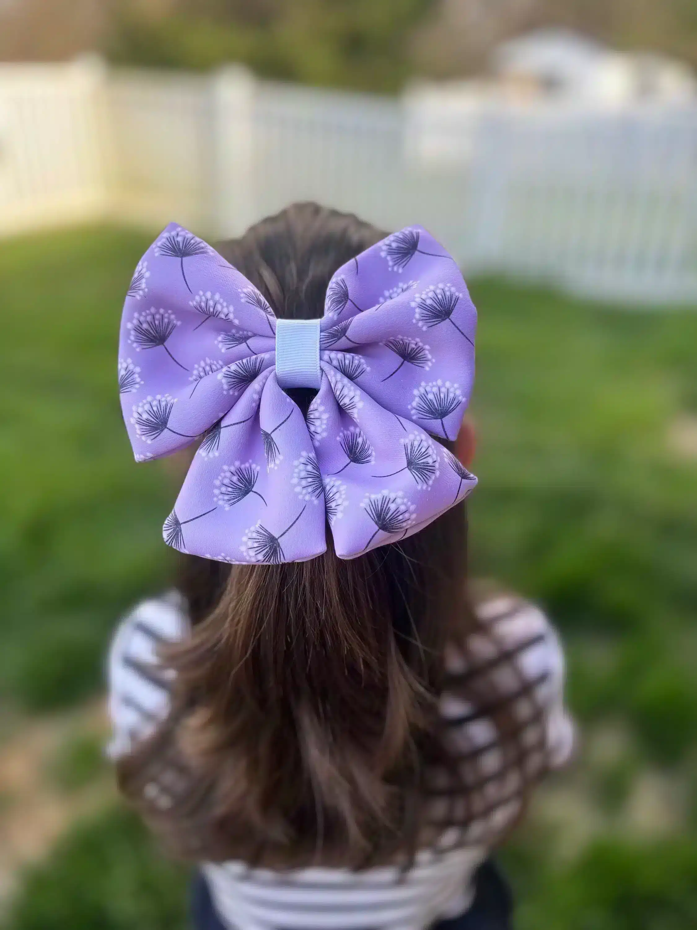 A handmade puffy hair bow with a dandelion print in lavender, worn by a girl, displayed against a green outdoor background. The bow features a unique, full design perfect for celebrating the Month of the Military Child.