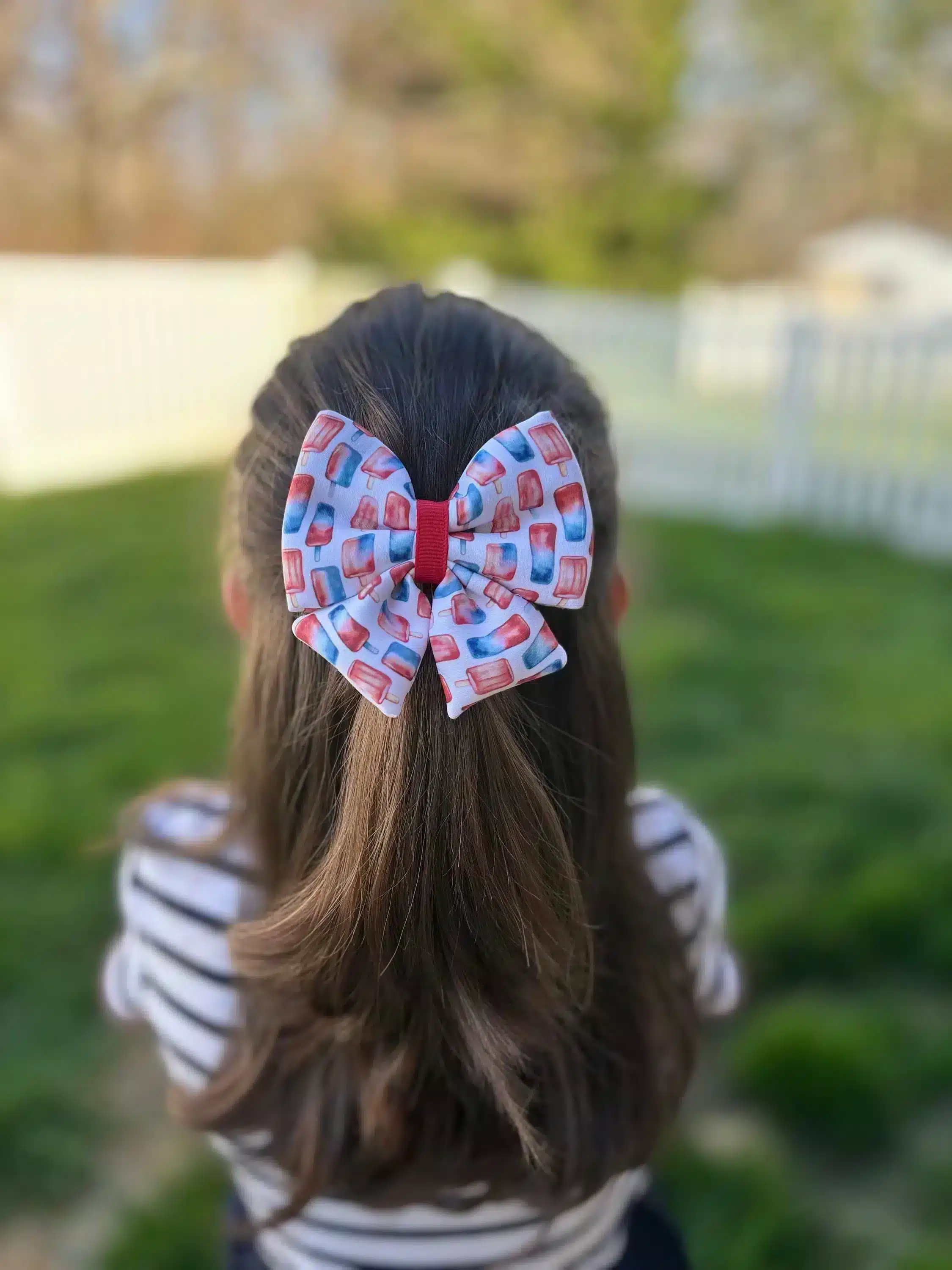 A handmade puffy hair bow with a patriotic popsicle print in red, white, and blue, worn by a girl, displayed against a green outdoor background. The bow features a unique, full design perfect for summer celebrations.