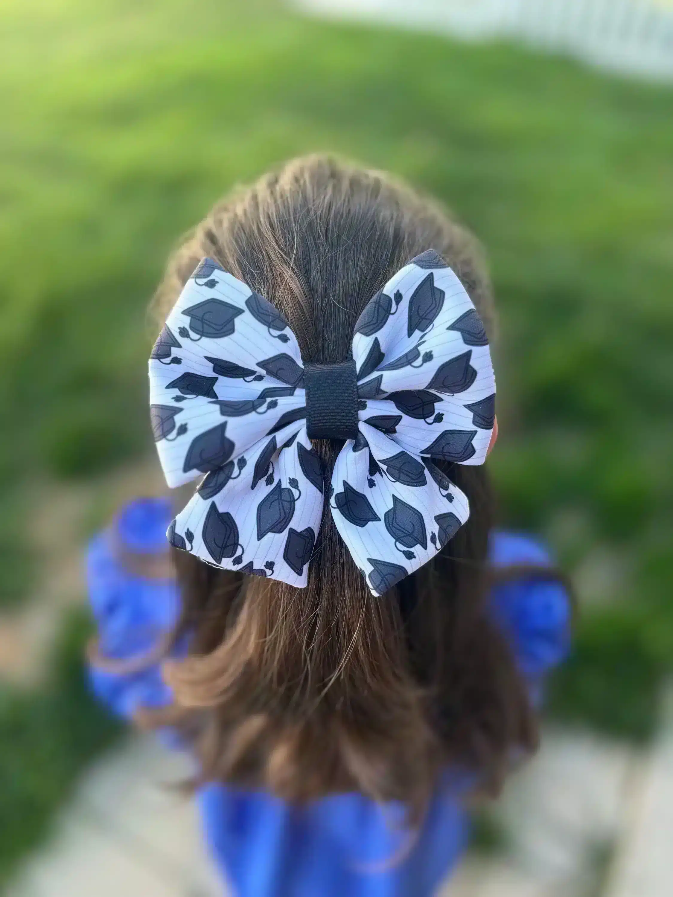 A handmade puffy hair bow with a graduation cap print, worn by a girl, displayed against a green outdoor background. The bow features a full, thick appearance, making it a standout accessory for graduation celebrations.