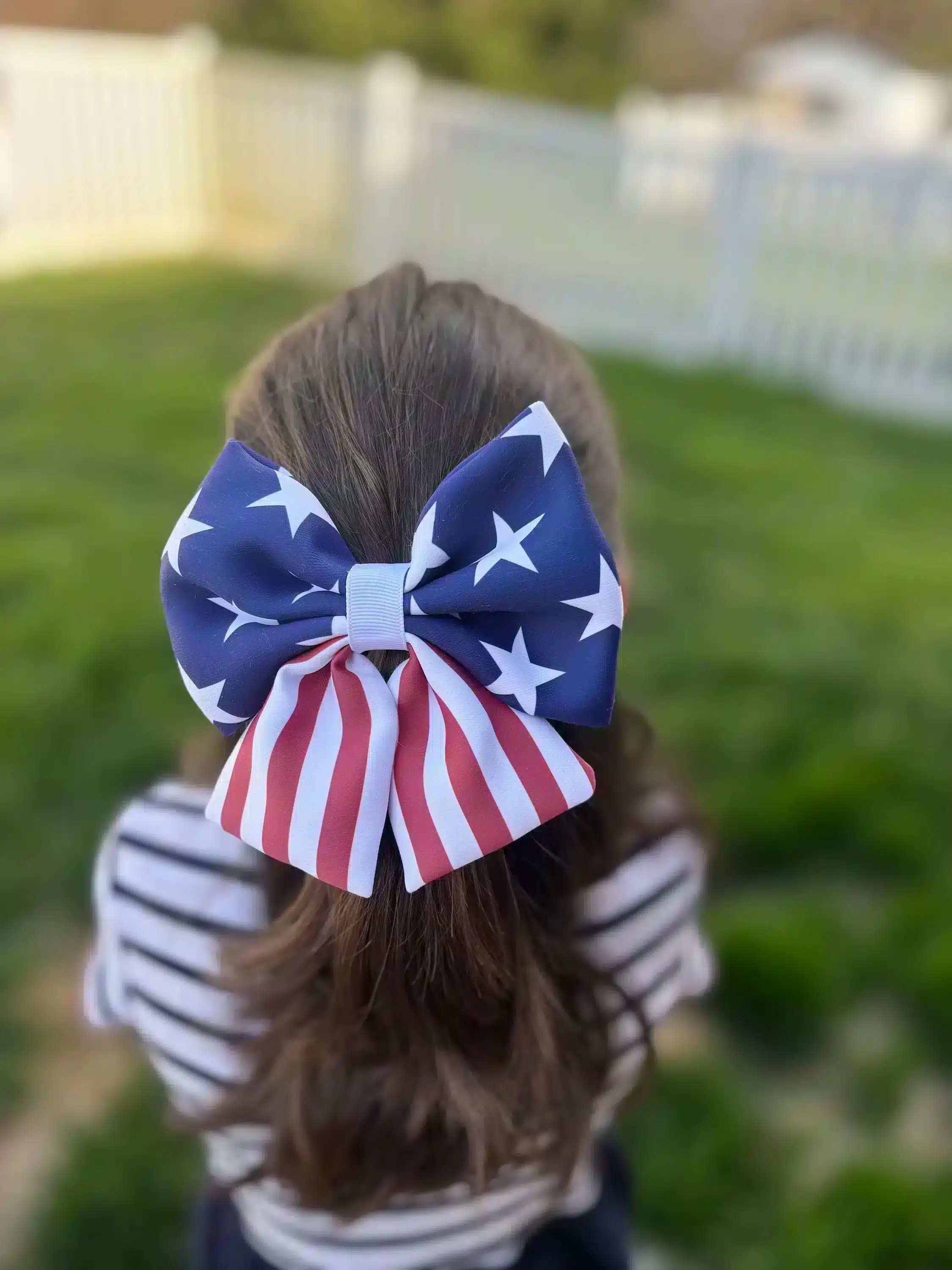 A handmade puffy hair bow with an American flag design, featuring stars and stripes, worn by a girl, displayed against a green outdoor background. The bow features a unique, full design perfect for patriotic celebrations.