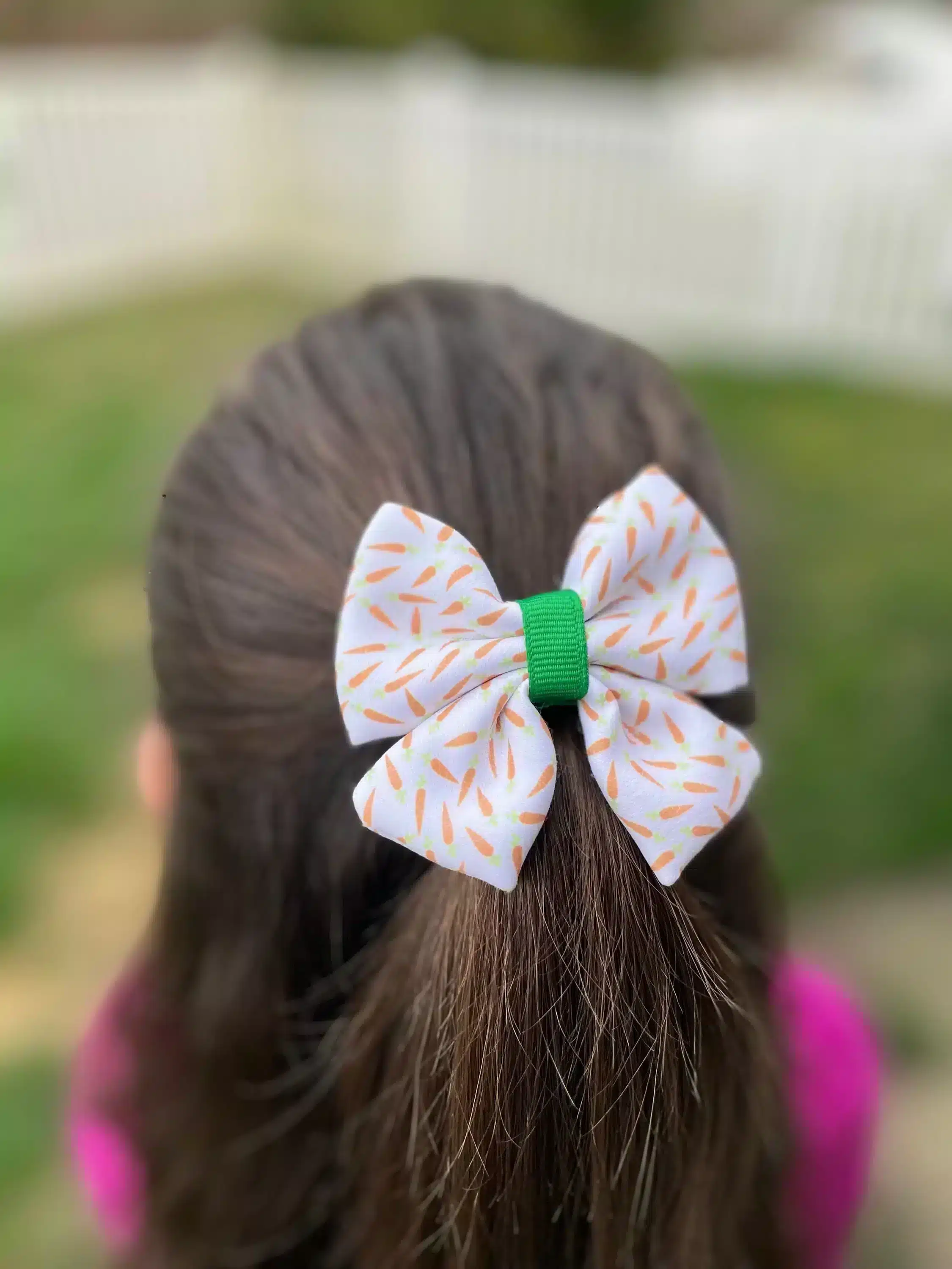 Back view of a girl wearing a white hair bow with a carrot print and green ribbon center in her brown hair.