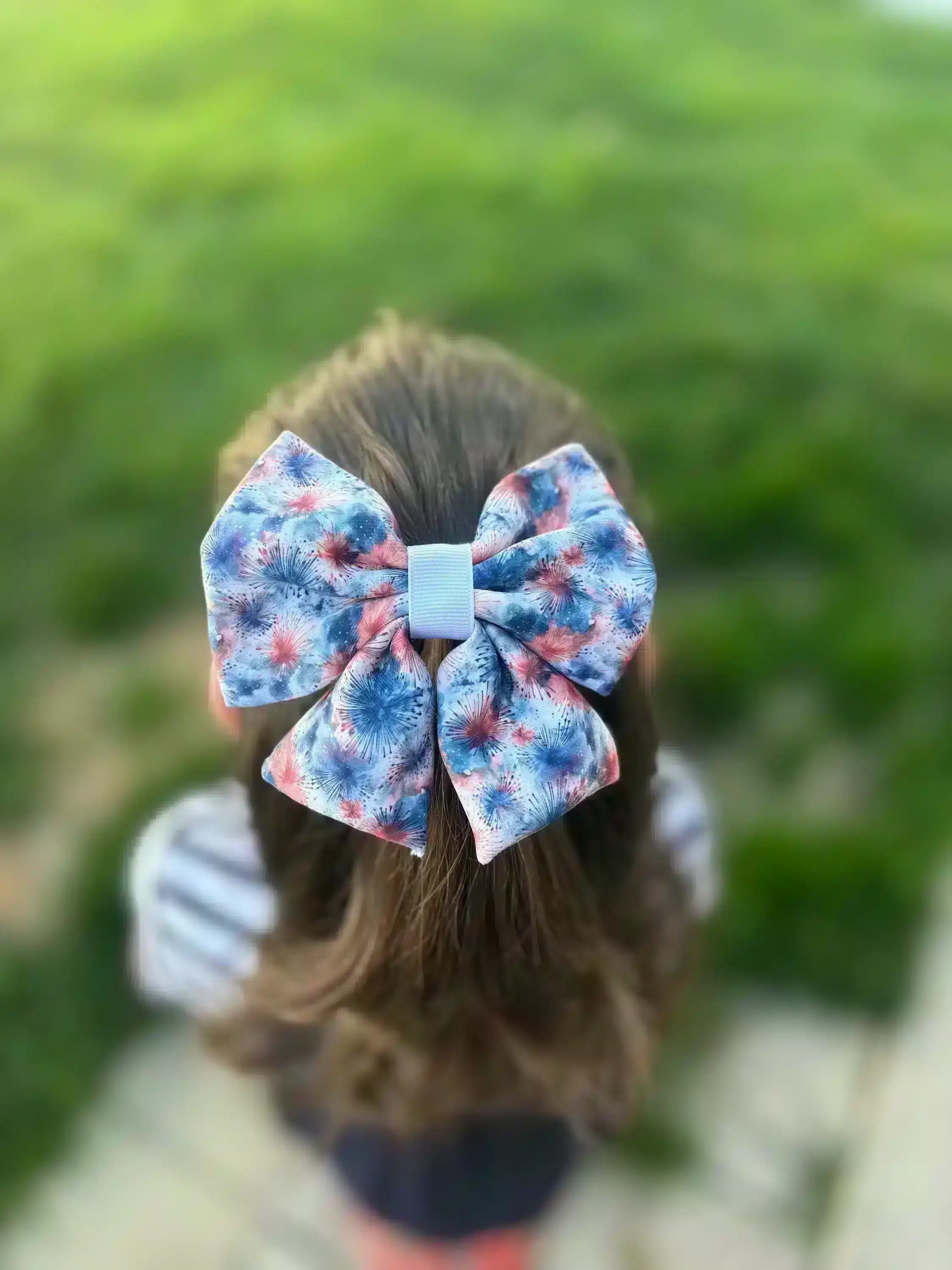 A handmade puffy hair bow with a firework print in red, white, and blue, worn by a girl, displayed against a green outdoor background. The bow features a unique, full appearance perfect for festive occasions.